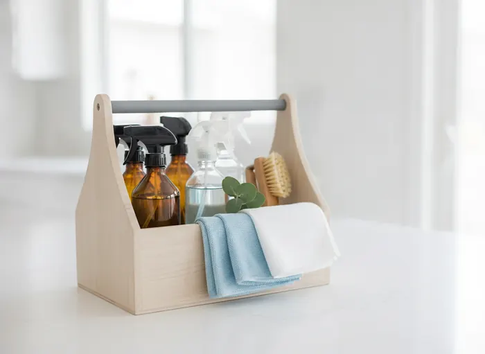 A wooden cleaning caddy with unlabeled glass spray bottles, pale blue microfiber cloths and a scrub brush